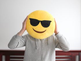 Rosto de emoji sorridente amarelo brilhante usando almofada de óculos escuros sustentada por uma pessoa Por iheartcreative