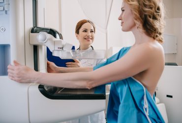 radiologista sorridente em pé perto do paciente enquanto faz diagnósticos de mamografia na máquina de raio-x Por LightFieldStudios