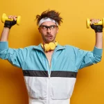 Jovem treina músculos das mãos com halteres, faz todos os esforços para levantar peso, faz reabilitação física, faz exercícios para os braços, posa em casa ginásio, vestido com roupas esportivas.
