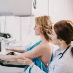 jovem radiologista em pé perto do paciente enquanto fazia o teste de mamografia na máquina de raio-x Por LightFieldStudios